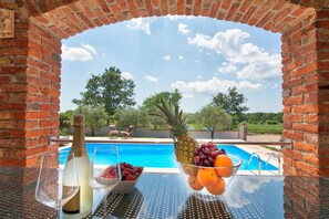 Et bord ved poolen med champagne, en skål med frugt og et glas vin, med udsigt over en swimmingpool og en klar himmel.
