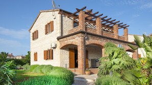 Ein zweistöckiges Haus mit Ziegelaußenwand, einer Holzpergola im oberen Bereich und einem Garten mit gepflegten Hecken.