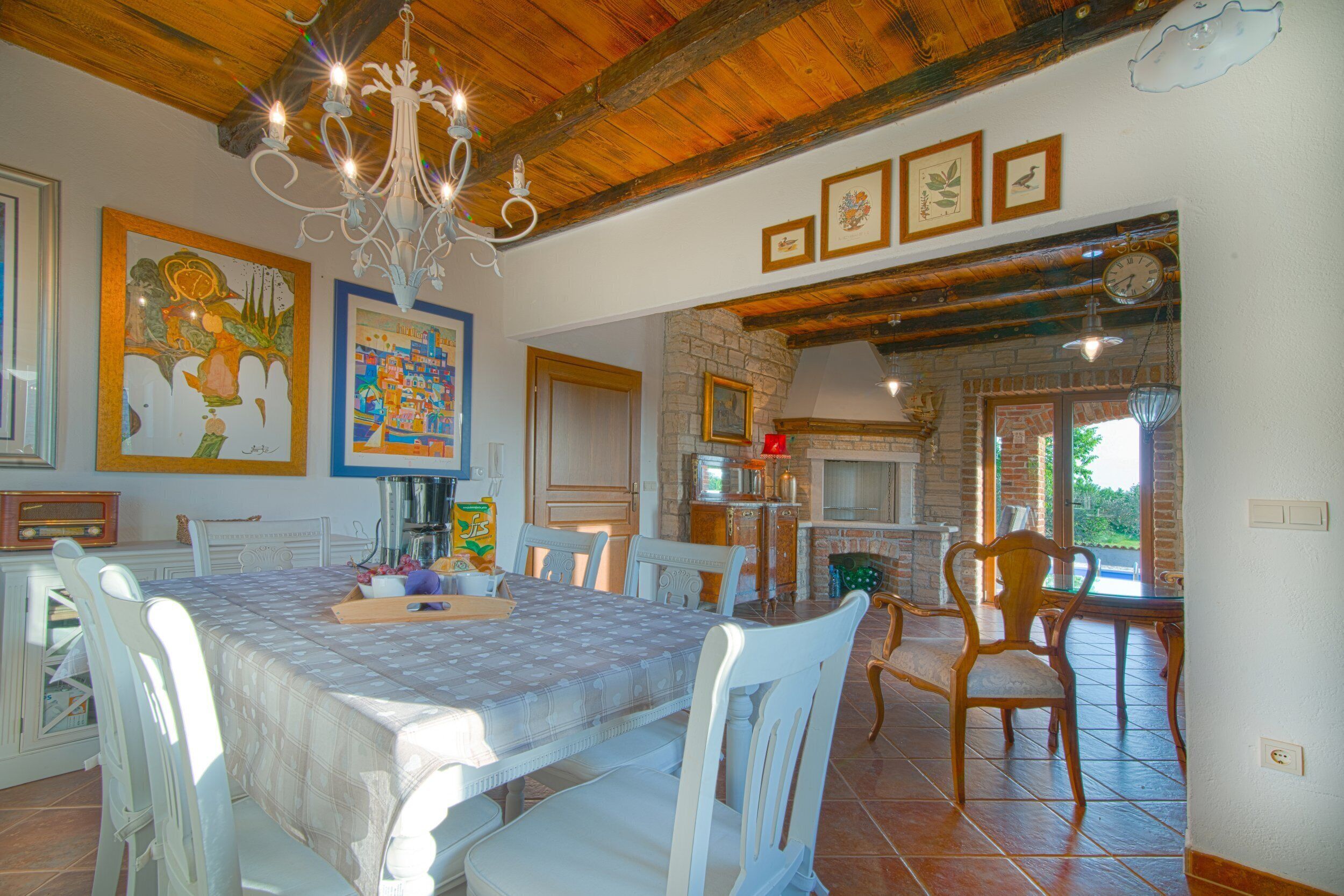 Un coin repas avec une grande table, des chaises blanches et un lustre. Les murs sont ornés de tableaux encadrés et une cheminée en briques est présente.