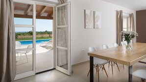 A room with a wooden table, white chairs, a vase with flowers, and a view of a swimming pool outside. - Villa Meli in Vodnjan (Vodnjan)