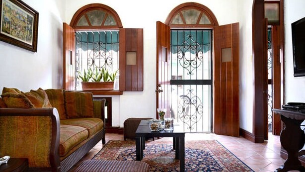 Un soggiorno con divano, tavolino e tappeto decorato. Due finestre presentano archi e inferriate in metallo.
