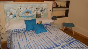 Un dormitorio con cama, almohadas, una mesita de noche, una lámpara, un taburete pequeño y una estantería en la pared con libros y objetos decorativos.