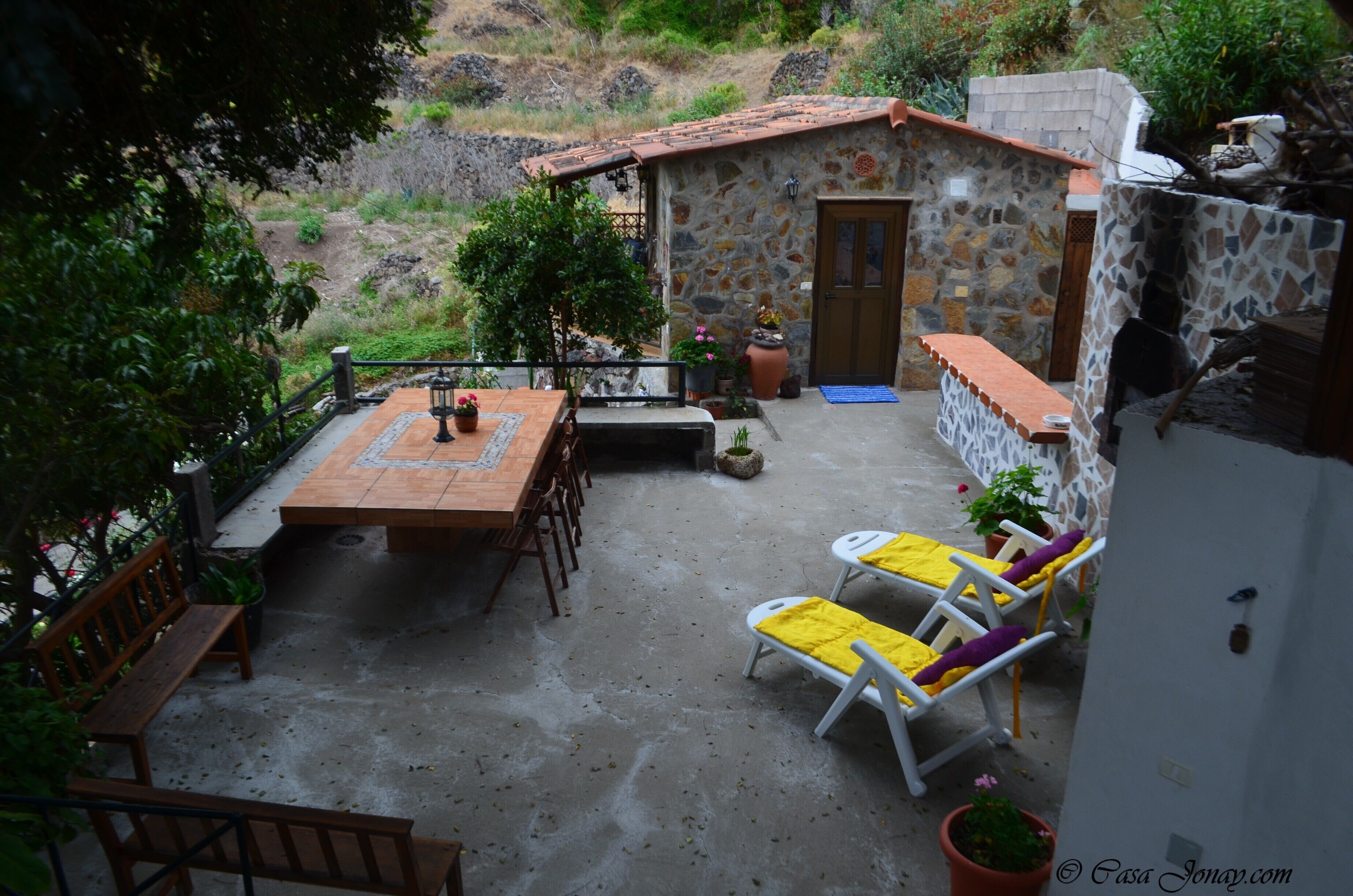 A stone building with a tiled roof, a wooden table, and two lounge chairs with yellow cushions.