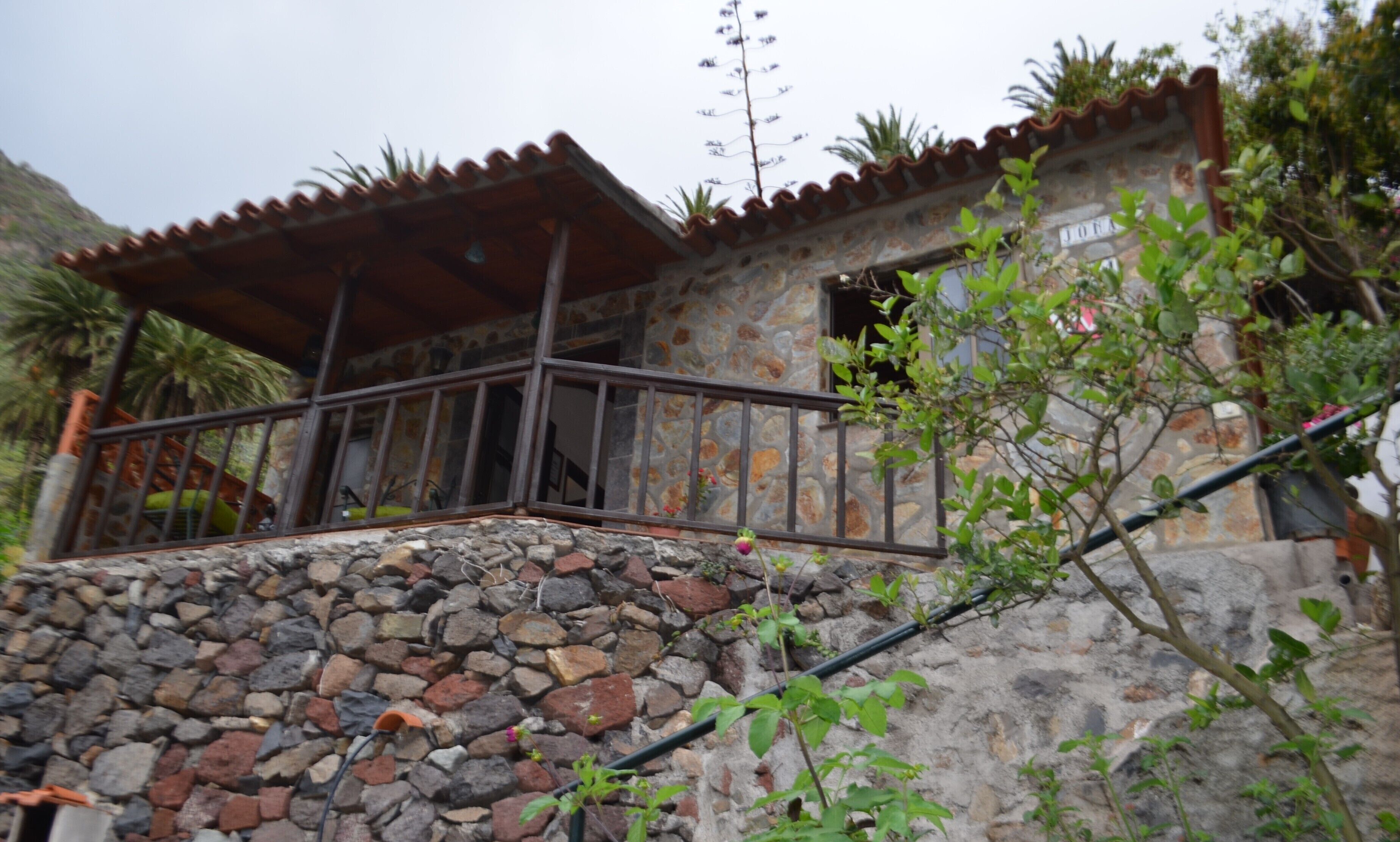 A stone house with a tiled roof, a balcony with metal railings, and a stone wall.