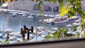 A marina with numerous boats docked, a stone building with arches in the background, and a view through a window with greenery in the foreground. - Apartment Marijana The most amazing breathtaking view to the sea and Old Town! (Dubrovnik)