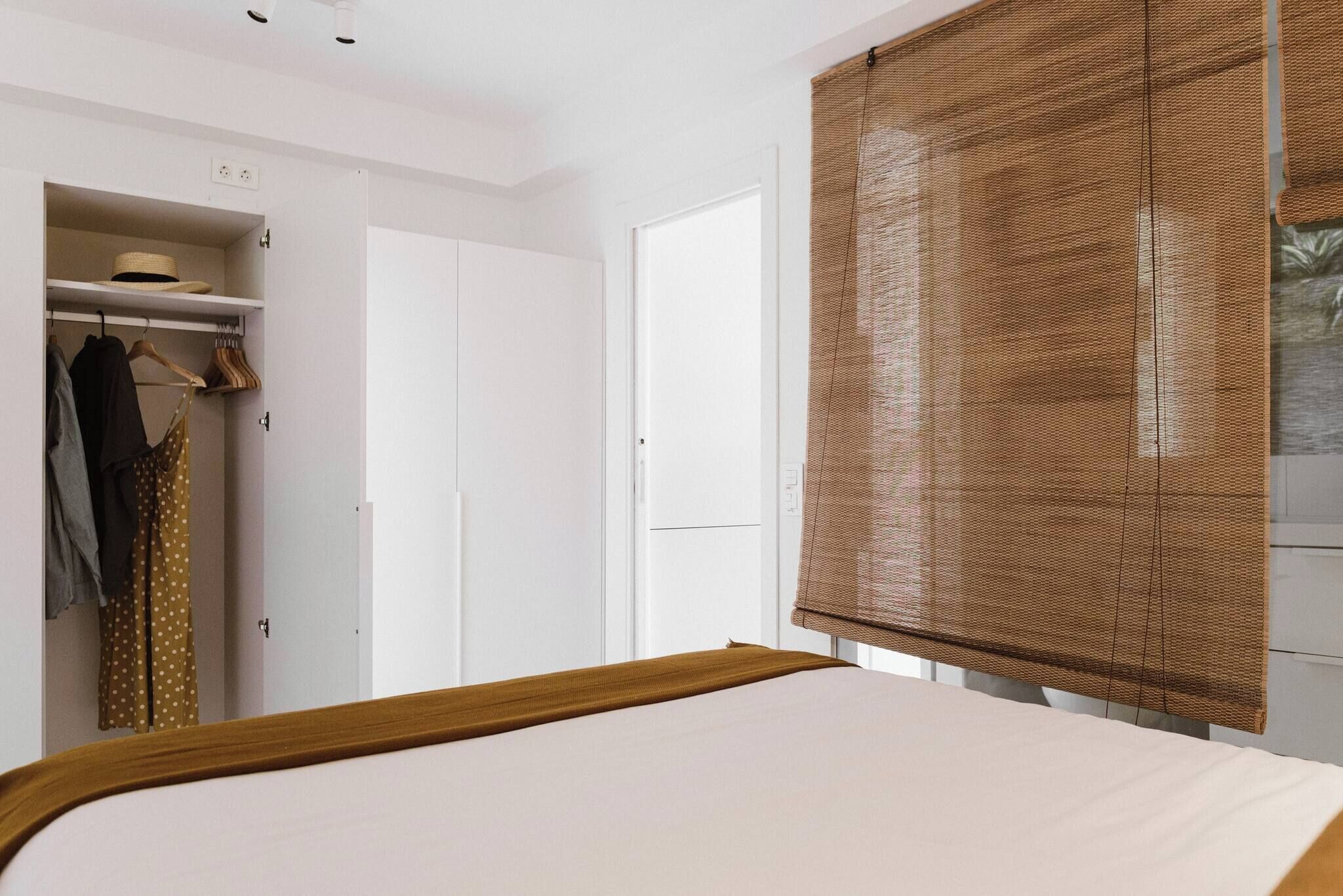 Un dormitorio con cama, armario, perchero y ventana con persiana.