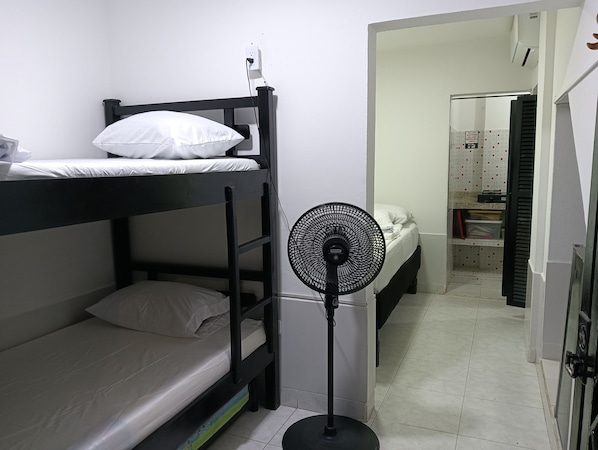 A dormitory room with bunk beds, a standing fan, and a small kitchenette area.
