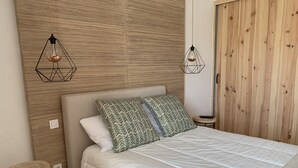 A bedroom with a bed, two geometric pendant lights, a wooden headboard, a small round table, and a wooden wardrobe.