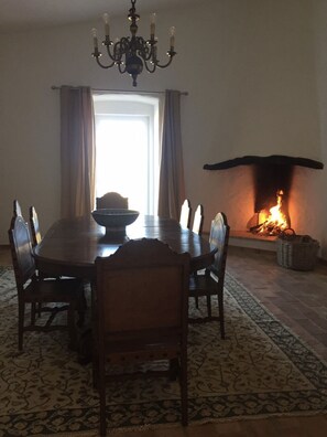 Une salle à manger avec une table et des chaises en bois, un lustre, une cheminée où brûle un feu et une fenêtre avec des rideaux.