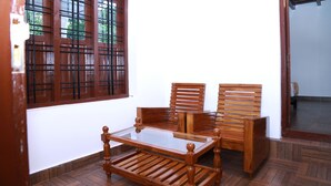 Een kamer met een houten tafel en twee stoelen, een raam met metalen tralies, en een houten deur.