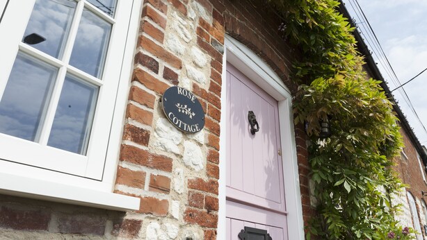 Um edifício de tijolos com uma janela branca e uma porta rosa. Uma placa na parede diz 'ROSE COTTAGE'.