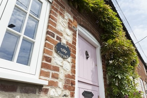 A brick building with a white window and a pink door. A sign on the wall reads "ROSE COTTAGE". - Rose Cottage -  a self catering cottage that sleeps 4 guests  in 2 bedrooms (Kings Lynn)