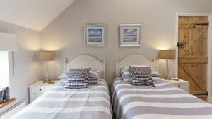 Quarto com duas camas, cada uma com roupa de cama listrada e almofadas xadrez. Há mesas de cabeceira com luminárias, uma porta de madeira e quadros nas paredes.