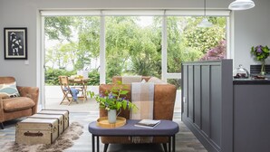 A modern living room with a brown leather sofa, a small table with a vase of flowers, and a kitchen area with a grey cabinet. - The Garden Rooms @ Drumagosker Rural Retreats (Limavady)