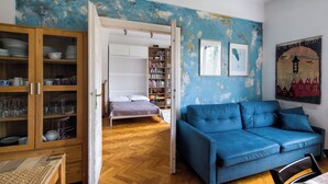 Une pièce avec un canapé bleu, un buffet en bois contenant de la vaisselle, un lit et une bibliothèque.