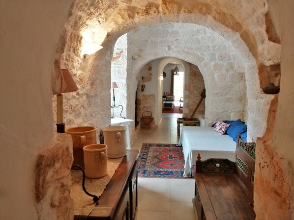 A rustic room with a stone archway, a bed with a patterned bedspread, a wooden bench, and a small table with a lamp.