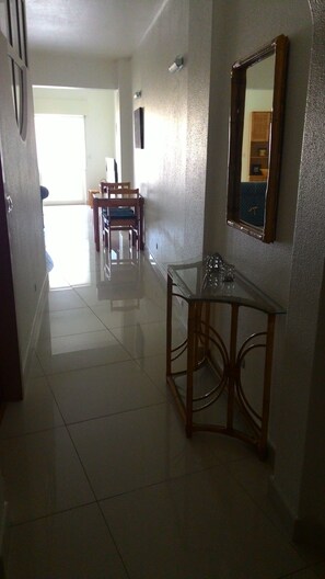 Um corredor com mesa e cadeiras de tampo de vidro, um espelho na parede e uma porta que dá para outro cômodo.