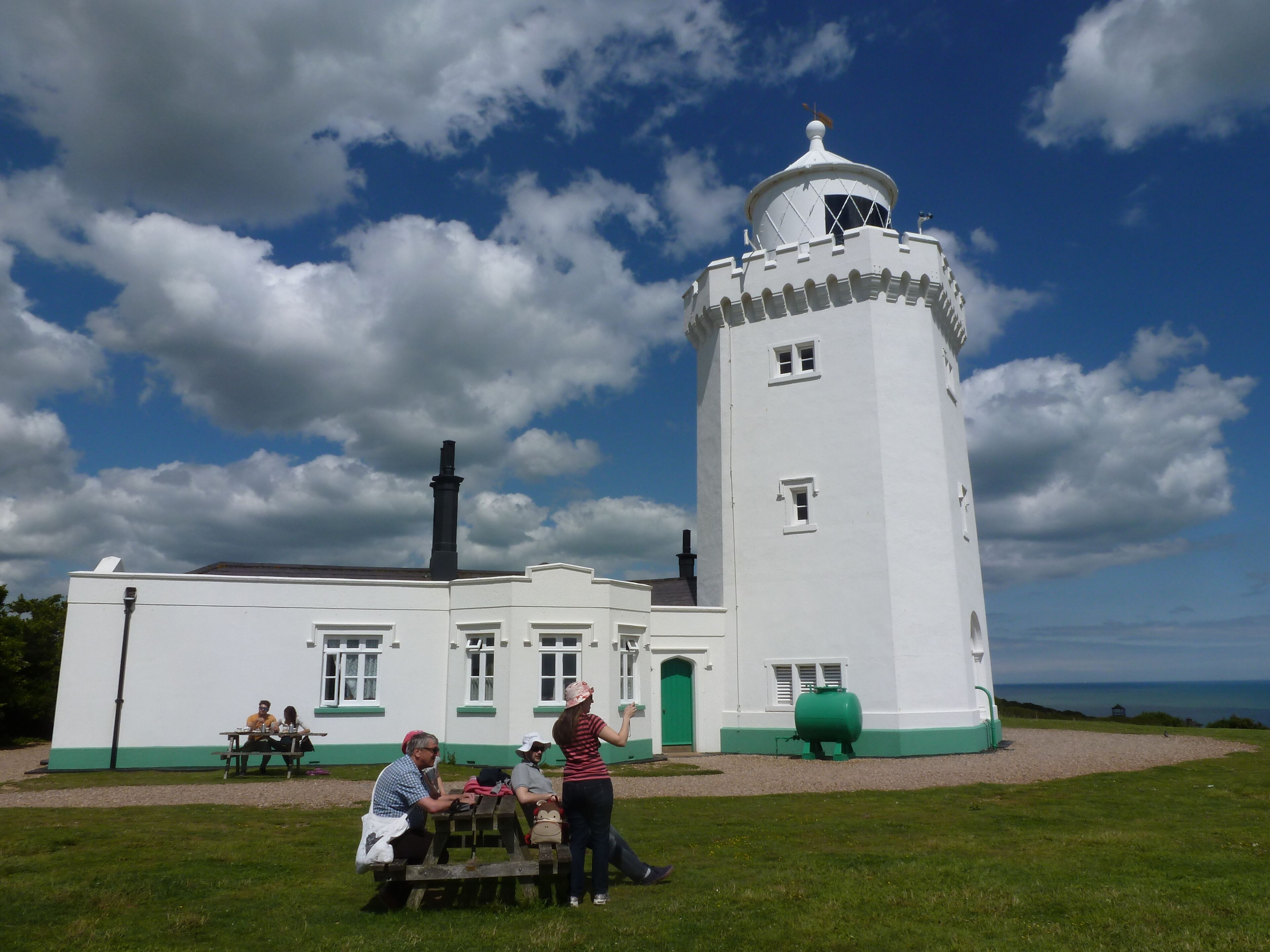 A white lighthouse with a green base, a small building with windows, and a grassy area with people.