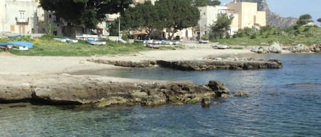 A coastal scene with clear water, rocky shore, boats, and buildings in the background.