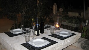 Ett utomhusbord med tillhörande stolar, dukat med tallrikar, bestick, glas och vinflaskor, omgivet av växter och en stenmur.