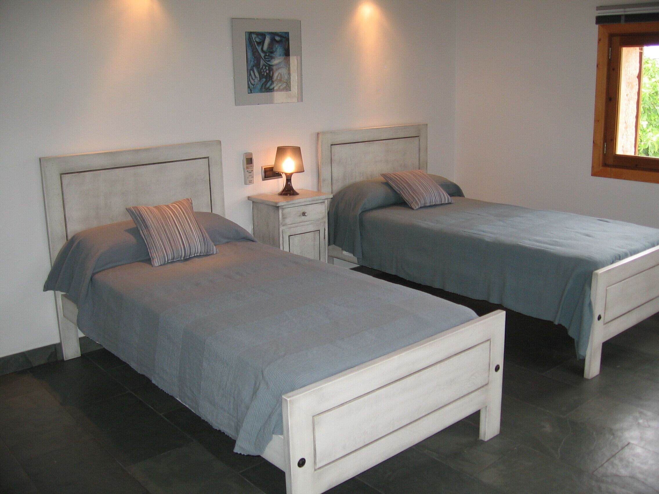 A room with two single beds, a nightstand with a lamp, a wall-mounted artwork, and a window with a view of greenery.
