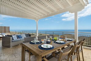 An outdoor dining area with a view of the sea, a wooden table set with plates and cutlery, and a wicker sofa with cushions. - ALÍA, modern house in village, magnificent sea and coastal views!  (Chania)
