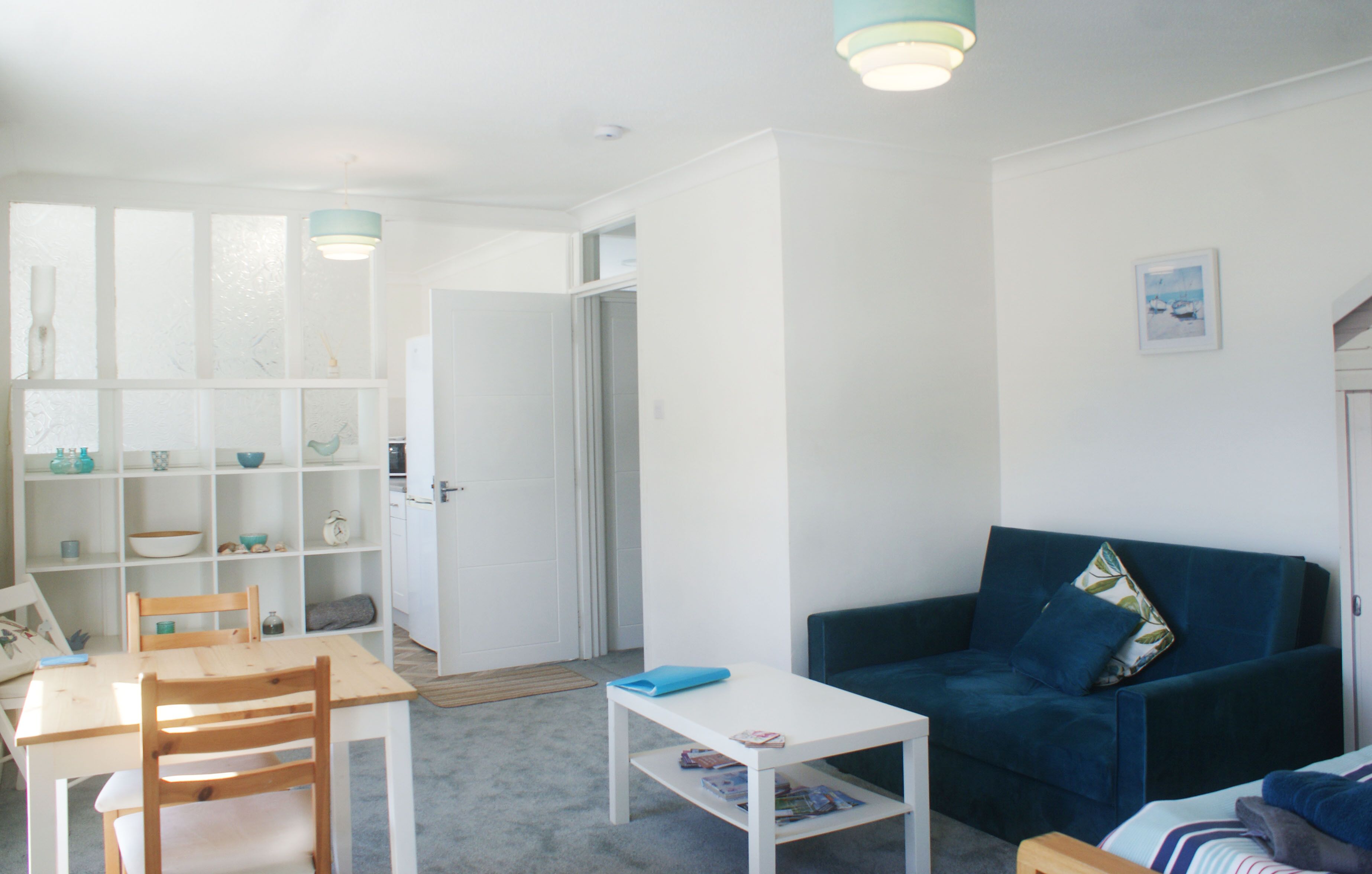 En lys stue med et hvidt bord, en blå sofa og en reol fyldt med pyngeting.
