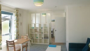 A room with a dining table, chairs, a blue sofa, and a white shelving unit with various items. - Central Sidmouth studio apartment with Sea Peeps (Sidmouth)
