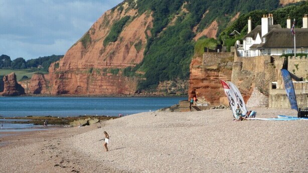 A beach with a rocky cliff, a thatched-roof building, and a flag on the ground. - Central Sidmouth studio apartment with Sea Peeps (Sidmouth)