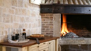 Une cheminée en pierre avec un feu de bois, un comptoir en bois sur lequel sont disposés un plateau de verres et des bouteilles, et une pizza.