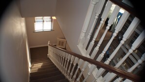 Una escalera con balaustres blancos y barandilla de madera, que conduce a un rellano con un cuadro enmarcado y una ventana con persianas.
