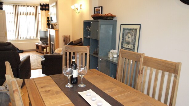 Comedor con una mesa de madera dispuesta para dos personas, una botella de vino y dos copas. Se observa un mueble azul con un cuadro en la parte superior, y un sofá de cuero negro al fondo.