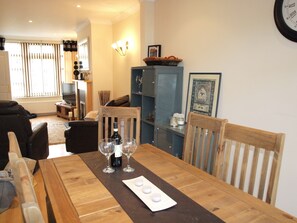 A dining area with a wooden table set for two, a wine bottle, and two wine glasses. There is a blue cabinet with a framed picture on top, and a black leather sofa in the background.