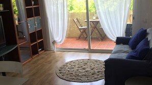 Una sala de estar con piso de madera, un sofá azul, un tapete redondo, una silla blanca y un puerta corrediza de vidrio que da a un patio exterior con una mesa y sillas.