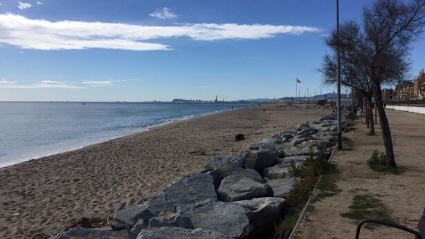 Una playa con un espigón rocoso, cielo despejado y edificios a lo lejos.