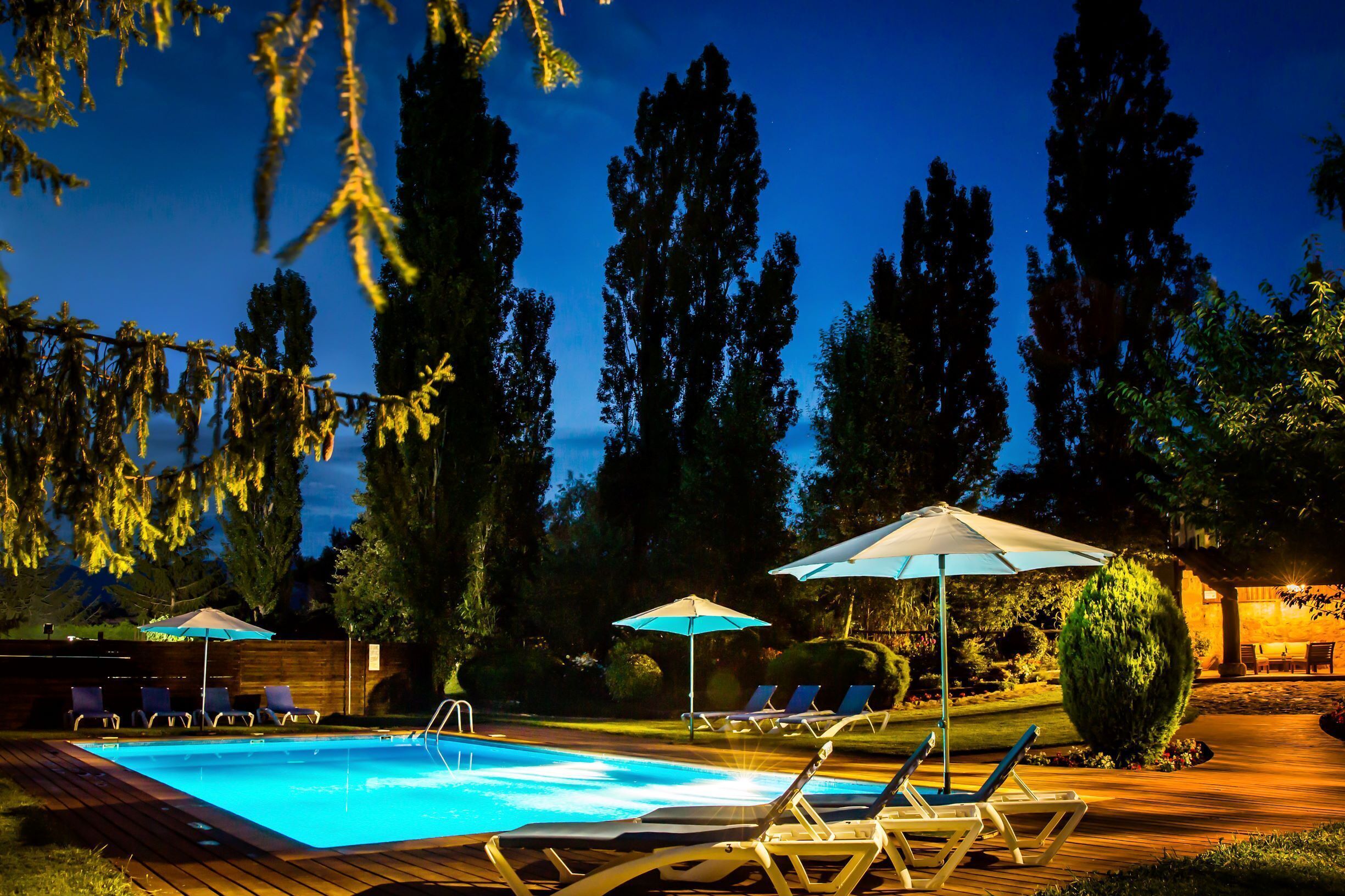 A swimming pool with lounge chairs and umbrellas, surrounded by tall trees and a wooden deck.