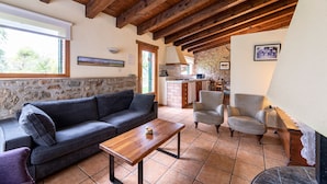 A cozy living room with a wooden ceiling, a stone wall, a sofa, a coffee table, and a kitchen area visible through an open doorway. - Casa Corral for 6 people (Lladurs)