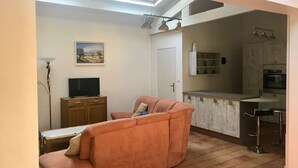 A living room with a sofa, a television, and a painting on the wall. There is a kitchen area with a countertop and a sink. - Villa with sea access on foot!  Quiet and large shady garden (Sanary-Sur-Mer)