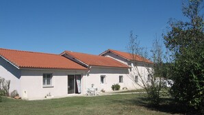 A two-story building with a red-tiled roof, a small patio with a table and chairs, and a grassy lawn.
