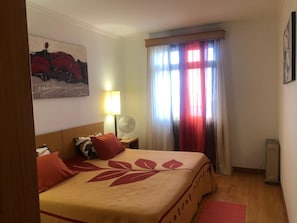 A hotel room with a bed, a nightstand, a lamp, a fan, and a window with curtains. - BARROS APARTMENT: Offers views of the ocean and mountains—visit Madeira’s many attractions. (Estreito De Câmara)