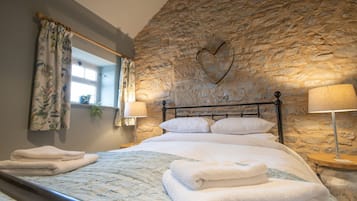 A bedroom with a stone wall, a heart-shaped decoration, a bed with white linens, two bedside lamps, and a window with floral curtains.