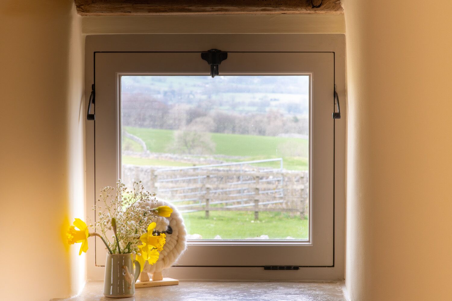 Une fenêtre donnant sur un paysage rural, un petit vase avec des fleurs et une sculpture en bois posée sur le rebord de la fenêtre.