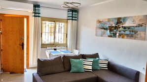Uma sala de estar com sofá, mesa e cadeiras, janela com cortinas e um quadro na parede.