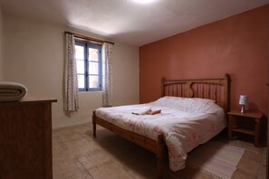 Una camera da letto con un letto in legno, una finestra con tende, un comodino con una lampada e un settimino in legno.
