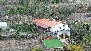 Una casa de una planta con techo rojo, un pequeño jardín delantero con césped verde y un muro de piedra al fondo.