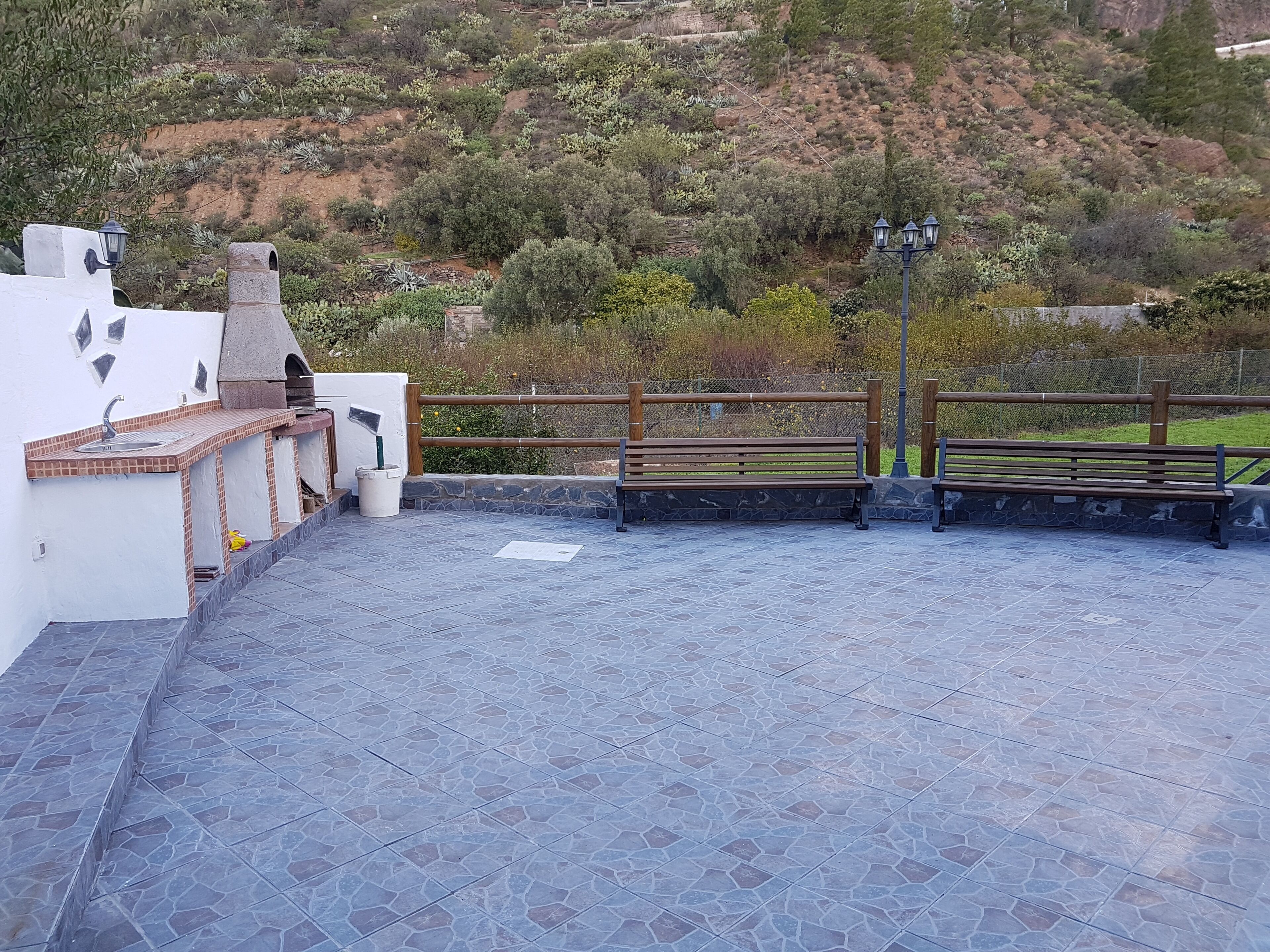 A rooftop terrace with a stone floor, a wooden bench, a metal railing, and a view of a hilly landscape.
