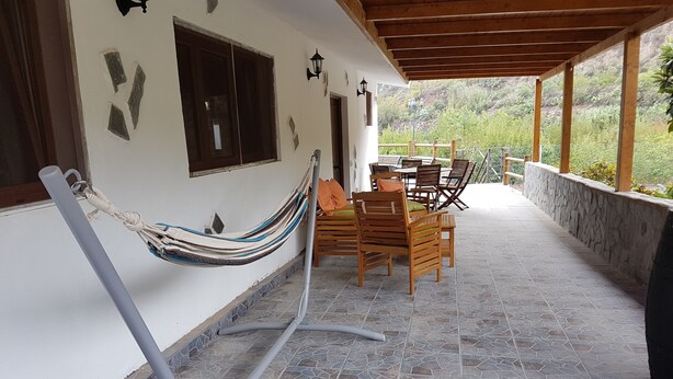 Un espacio de patio techado con una hamaca, sillas de madera y una mesa.