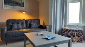A modern living room with a grey sofa, a small table with decorative items, a lamp, and a window with curtains. - Bute House - Great Sea Views! (Rothesay)