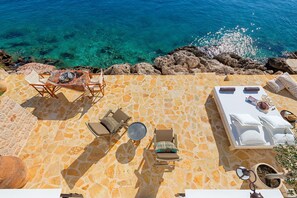 A sunlit outdoor seating area with stone flooring, lounge chairs, and a table, overlooking a clear blue sea.