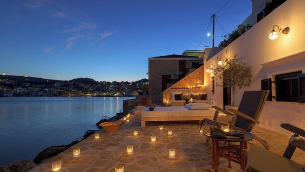 A stone-floored outdoor seating area with candles, a bed, and lounge chairs overlooking a calm body of water at dusk. - Halki Sea House, Luxury villa by the sea (HALKI ISLAND)